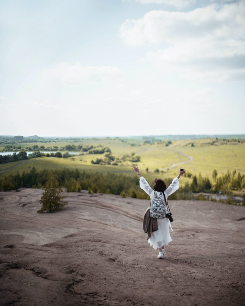 a boho-looking woman with her hands up in the air, overlooking a green landscape